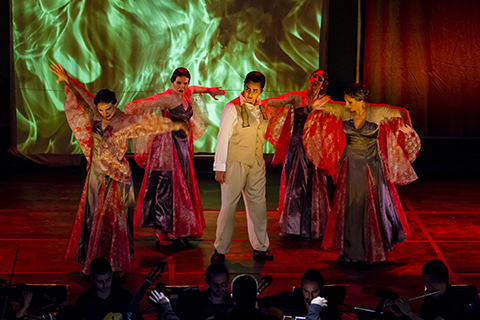 Five actors perform a scene from an opera on stage while an orchestra plays below them