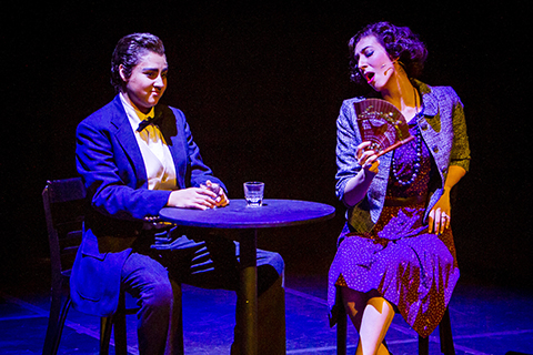 Two performers are sitting at a table in a scene from an opera