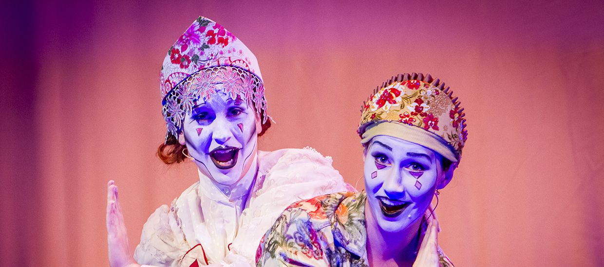 Two opera singers in expressive makeup smile as they perform on stage